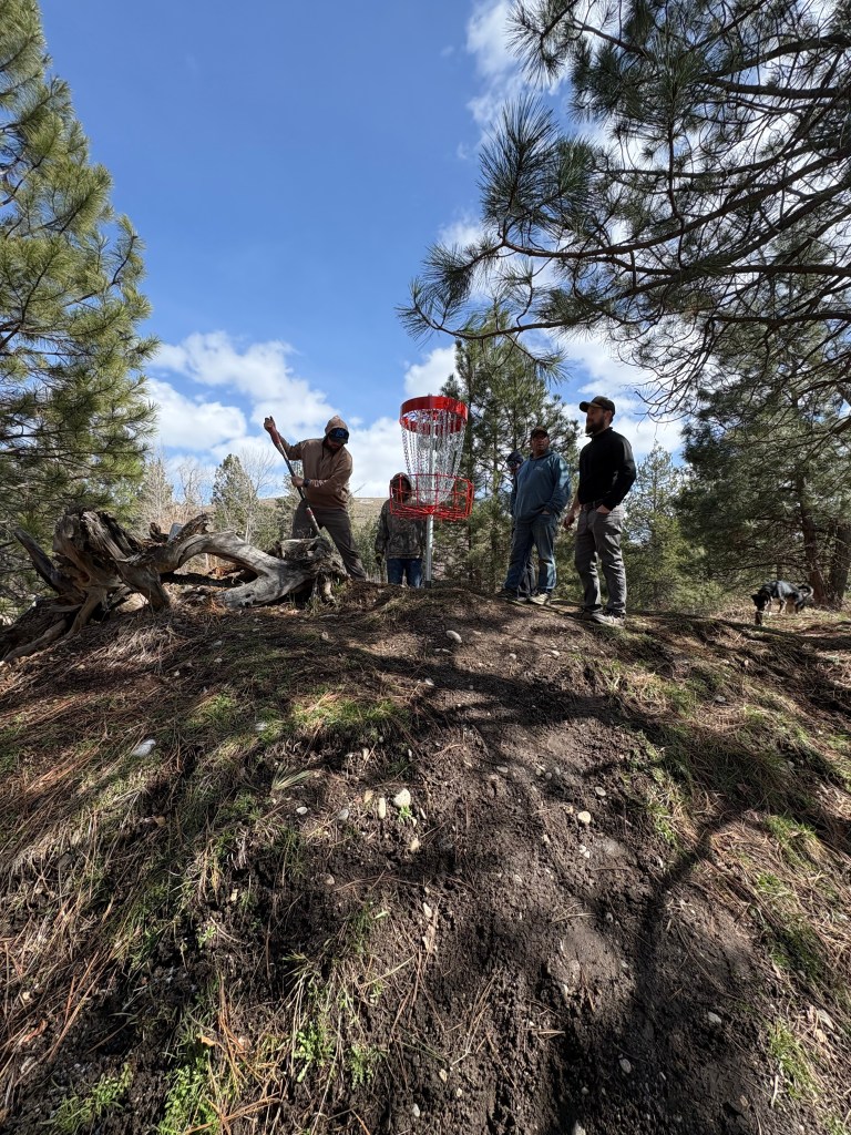 Group of people installing a frisbee golf basket om top of a hill at the Darby disc Golf course with a nice sunny day background