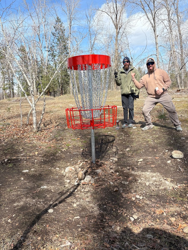 Two guys standing behind an Orange Disc Golf Basket on a nice sunny day.